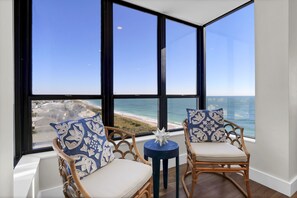 A room with two wicker chairs with white cushions and blue patterned pillows, a small round table with a white object, and a large window offering a view of the beach and ocean. - Station One 8A - Upscale 3 bedroom/2 bathroom oceanfront penthouse. (Wrightsville Beach)