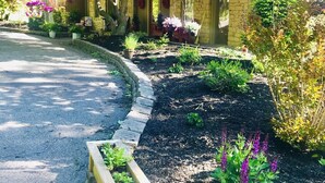 Una propiedad residencial con fachada de piedra, camino de entrada y jardín con diversas plantas y flores.
