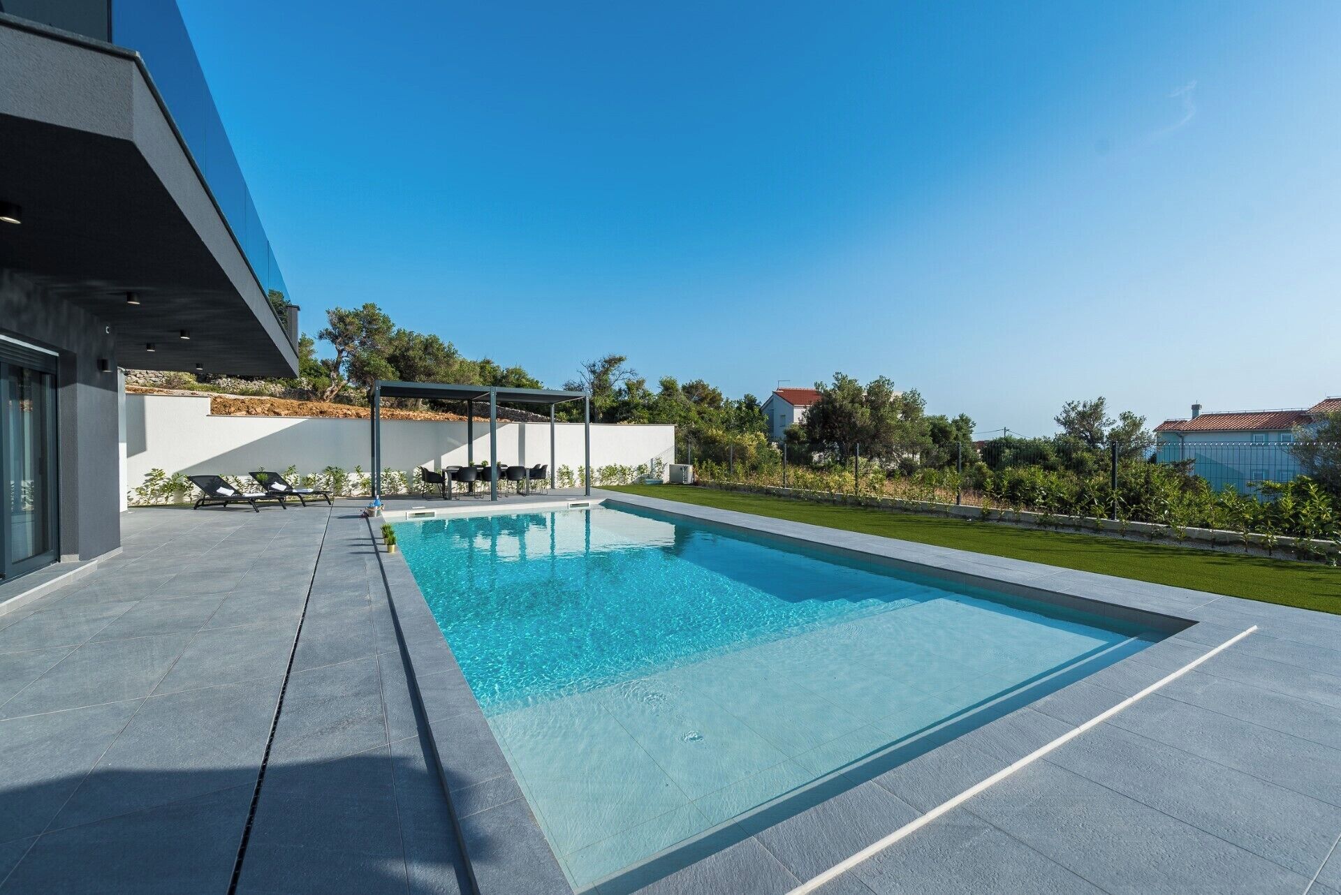 A modern outdoor swimming pool with a covered seating area, surrounded by a well-maintained lawn and a clear blue sky.