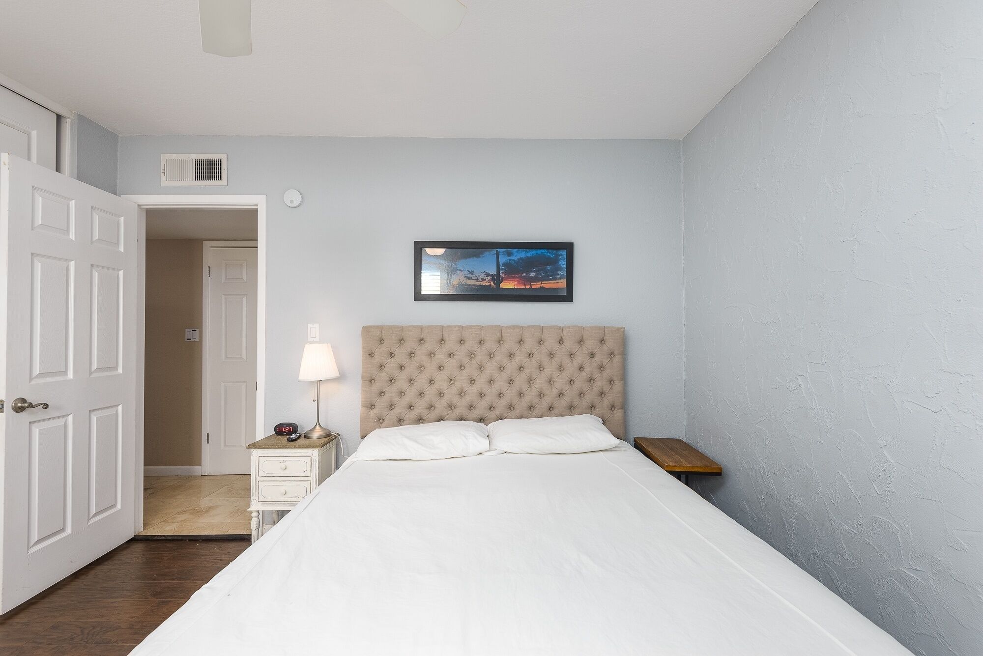 A bedroom with a tufted headboard, a bedside table with a lamp, a framed picture on the wall, and a door leading to another room.
