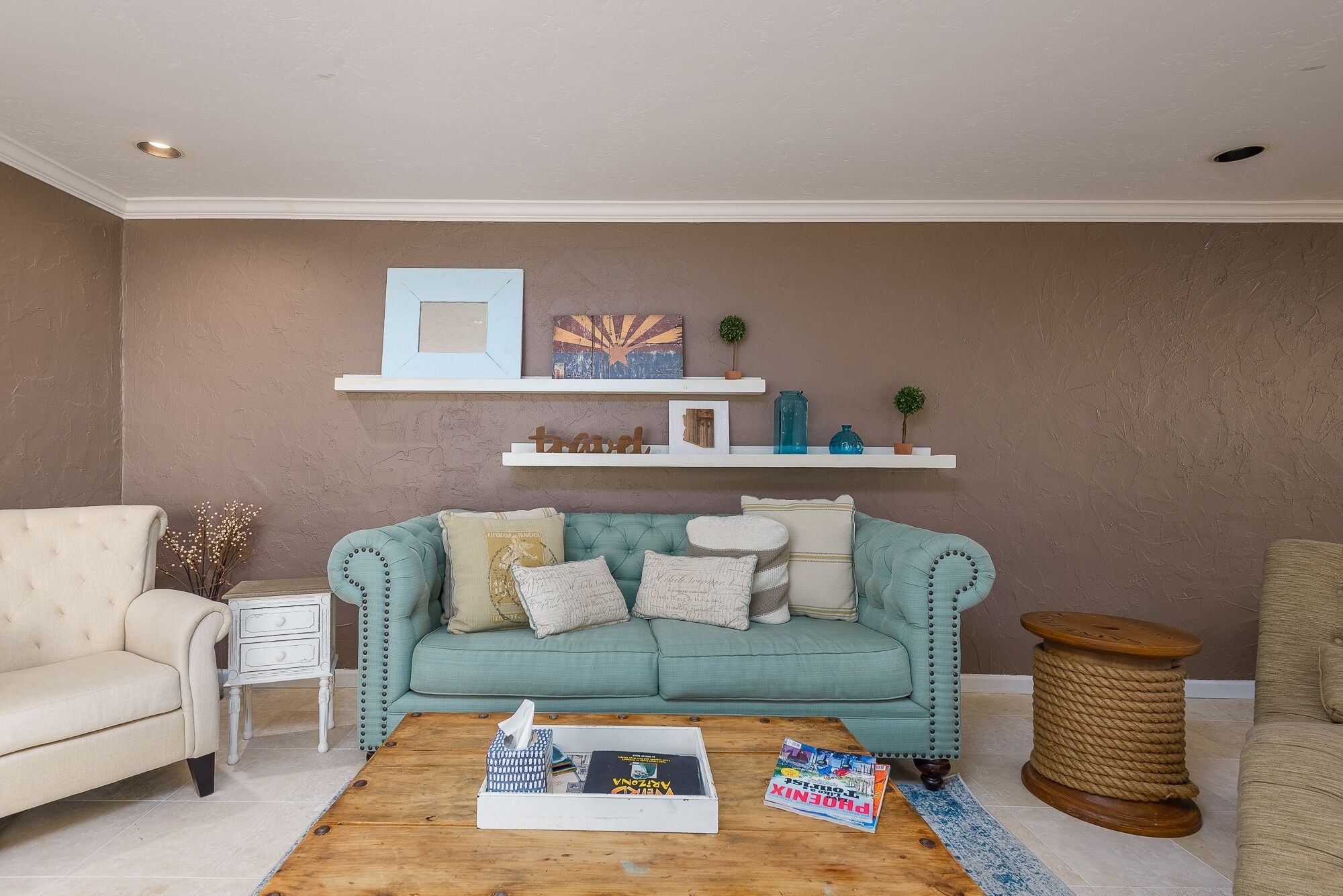 Una sala de estar con un sofá color turquesa, un sillón color beige, una mesa de centro de madera y una mesita auxiliar blanca.