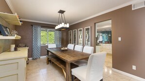 A dining area with a wooden table, white chairs, a chandelier, and a window with patterned curtains.