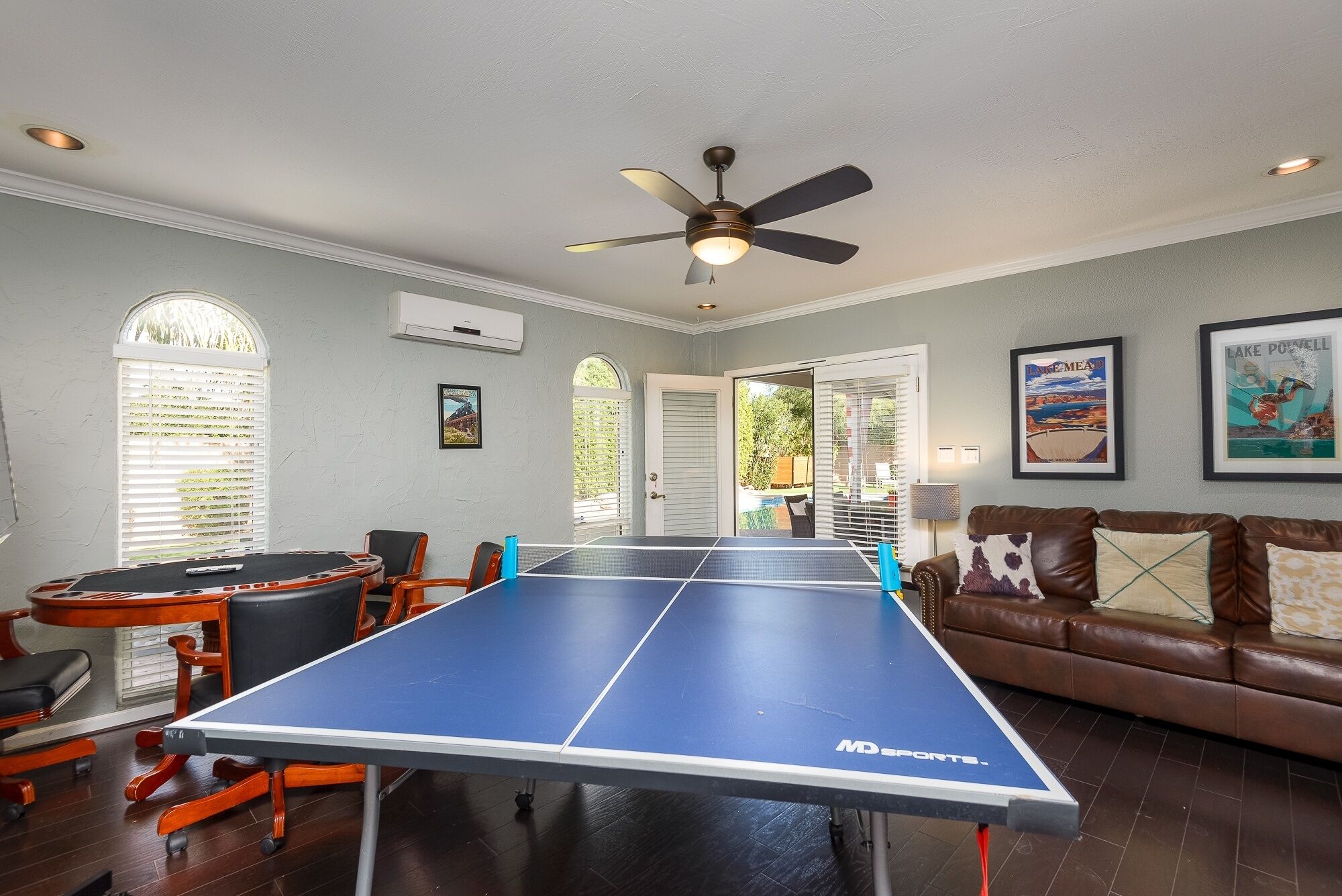 A ping pong table in a room with a ceiling fan, a brown leather sofa, and framed artwork on the walls.