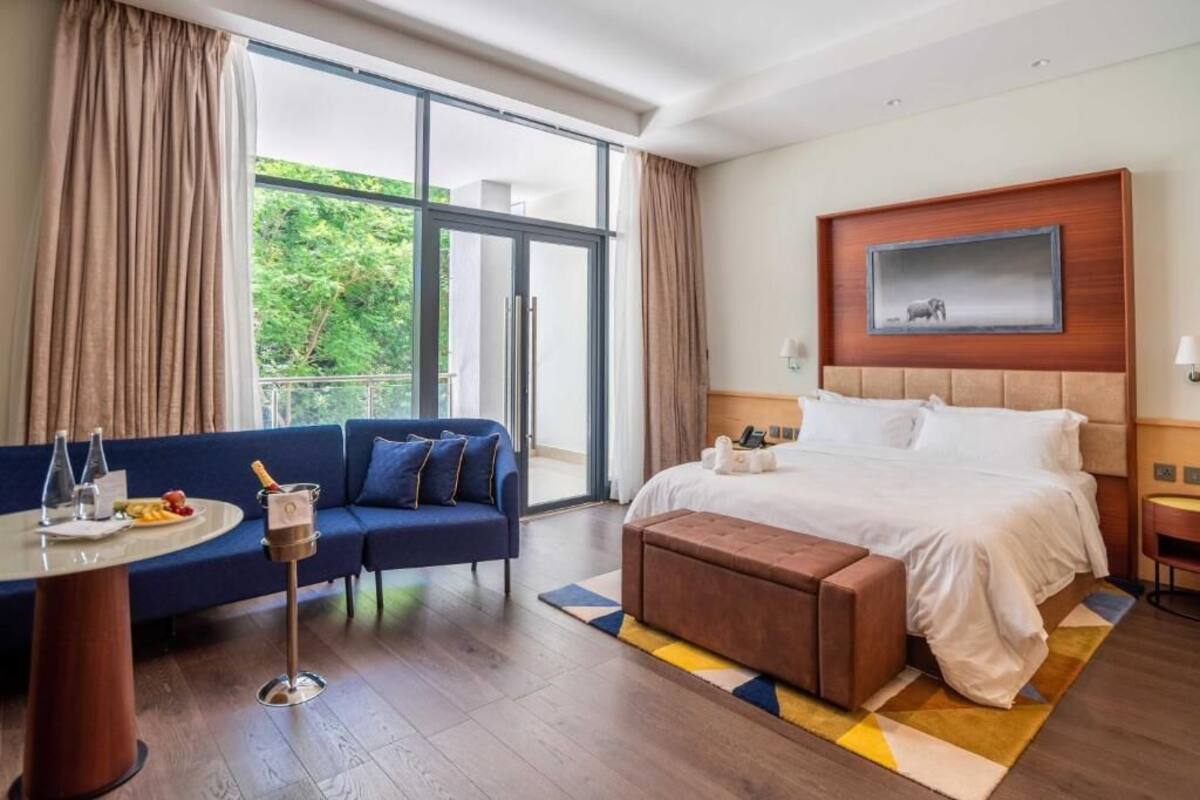 A hotel room with a large bed, a sofa, a small table with a drink, and a view of greenery outside.
