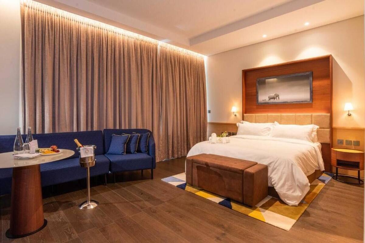 A hotel room with a bed, a sofa, a small table with a bottle and glasses, and a large window with curtains.