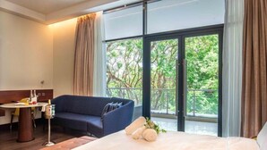 A hotel room with a bed, a sofa, a small table with a lamp, and a window with a view of trees. - The Quorum (Lusaka)