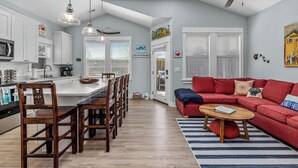 A modern kitchen with white cabinets, a wooden dining table, and pendant lights. There is a red sofa and a coffee table in the living area.