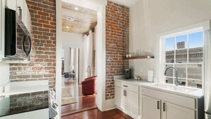 A kitchen with white cabinets, a brick accent wall, a window with a view, and a red chair in the dining area.
