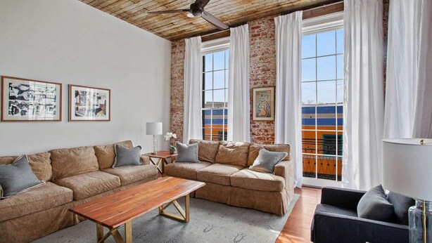 A living room with a wooden ceiling, a brick wall, and large windows with white curtains. There is a sofa set, a coffee table, and a lamp.