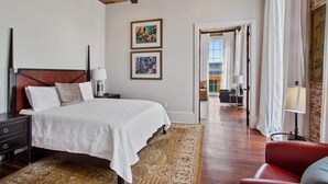 A bedroom with a wooden ceiling, a bed with white bedding, two framed artworks, a nightstand, a red chair, and a view through an open door to a balcony.