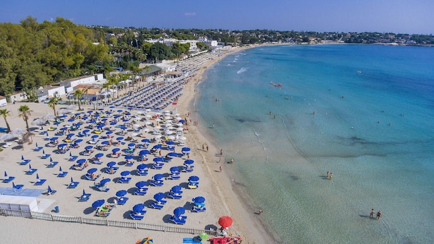 A beach with rows of blue umbrellas and sun loungers, a clear blue sea, and a line of trees and buildings in the background. - Futura Club Spiagge Bianche (Syracuse)