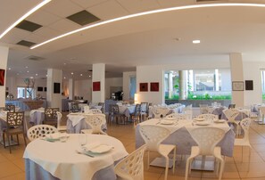 A spacious dining area with round tables and white chairs, featuring a modern design with recessed lighting and large windows offering a view of greenery outside. - Futura Club Spiagge Bianche (Syracuse)
