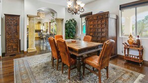 A dining area with a wooden table and chairs, a chandelier, and a large window with a view. - Luxury 7BR Golf Mansion: Pool, Spa, Tennis Court (Scottsdale)