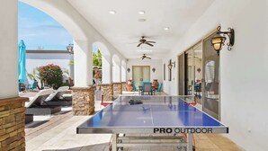 An outdoor ping pong table labeled "PRO OUTDOOR" in a covered patio area with stone pillars, lounge chairs, and potted plants. - Luxury 7BR Golf Mansion: Pool, Spa, Tennis Court (Scottsdale)