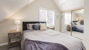 A bedroom with a large bed, a nightstand with a lamp, a window with blinds, and a mirror.