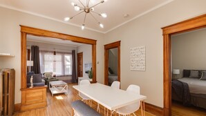 A spacious room with a wooden floor, a white dining table with chairs, a bench, and a bed in the adjacent room.