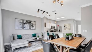 Una sala moderna con sofá, mesa de centro y mesa de comedor con sillas. Se ve una cocina al fondo con armarios y electrodomésticos.
