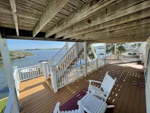 A wooden deck with white railings, a hammock, and two white chairs. A staircase leads to a covered area with a view of the water.