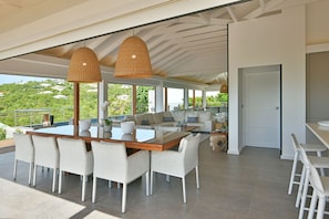 A modern outdoor dining area with a wooden table and white chairs, large wicker pendant lights, and a view of lush greenery. - Villa Avalon  | Ocean View - Located in  Wonderful Gouverneur with Private Pool (Gouverneur)