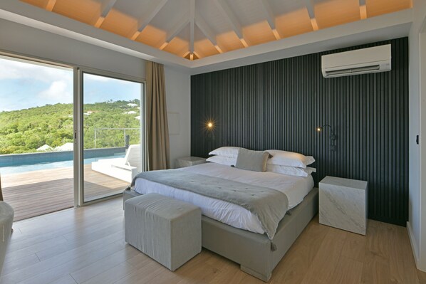 A modern bedroom with a large bed, a view of a pool and greenery, and an air conditioning unit on the wall.