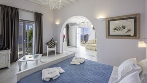 Et moderne hotelværelse med en stor seng, et siddeområde med en hvid stol, et lille bord og havudsigt gennem en  åben dør.