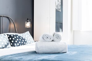 Un lit bien fait, avec des serviettes blanches pliées en rouleaux, un coussin à pois et une lampe de chevet.