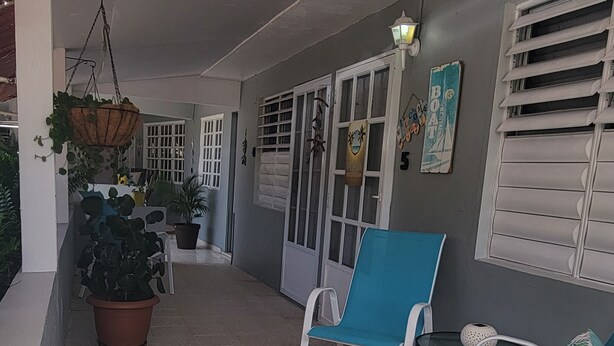 A covered outdoor seating area with chairs and tables, potted plants, and a wall with a door and window. - Playa Escondida (Fajardo)