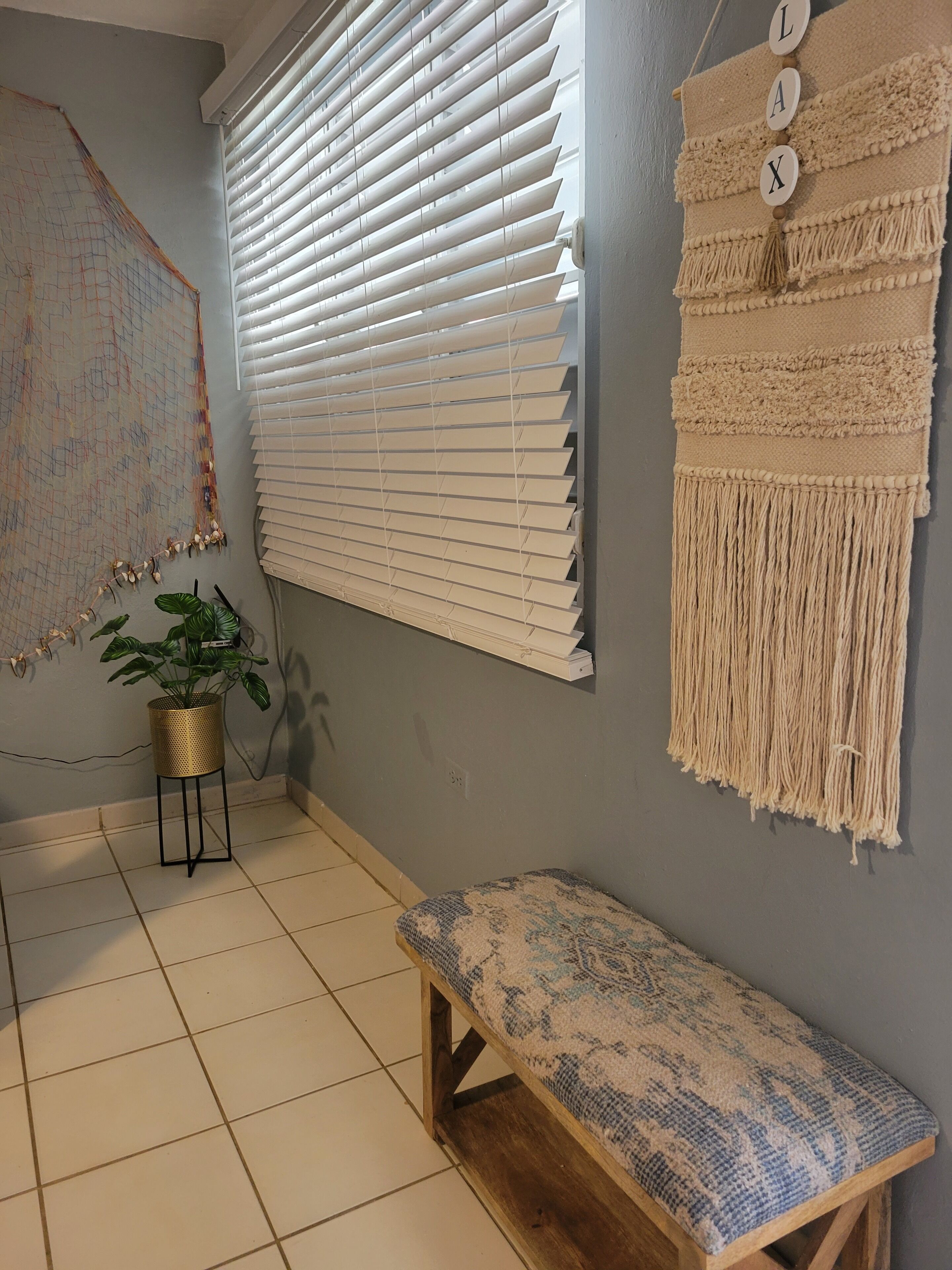 Una habitación con un ventanal grande cubierto por persianas, un colgador de macramé en la pared, un banco estampado y una planta en maceta.