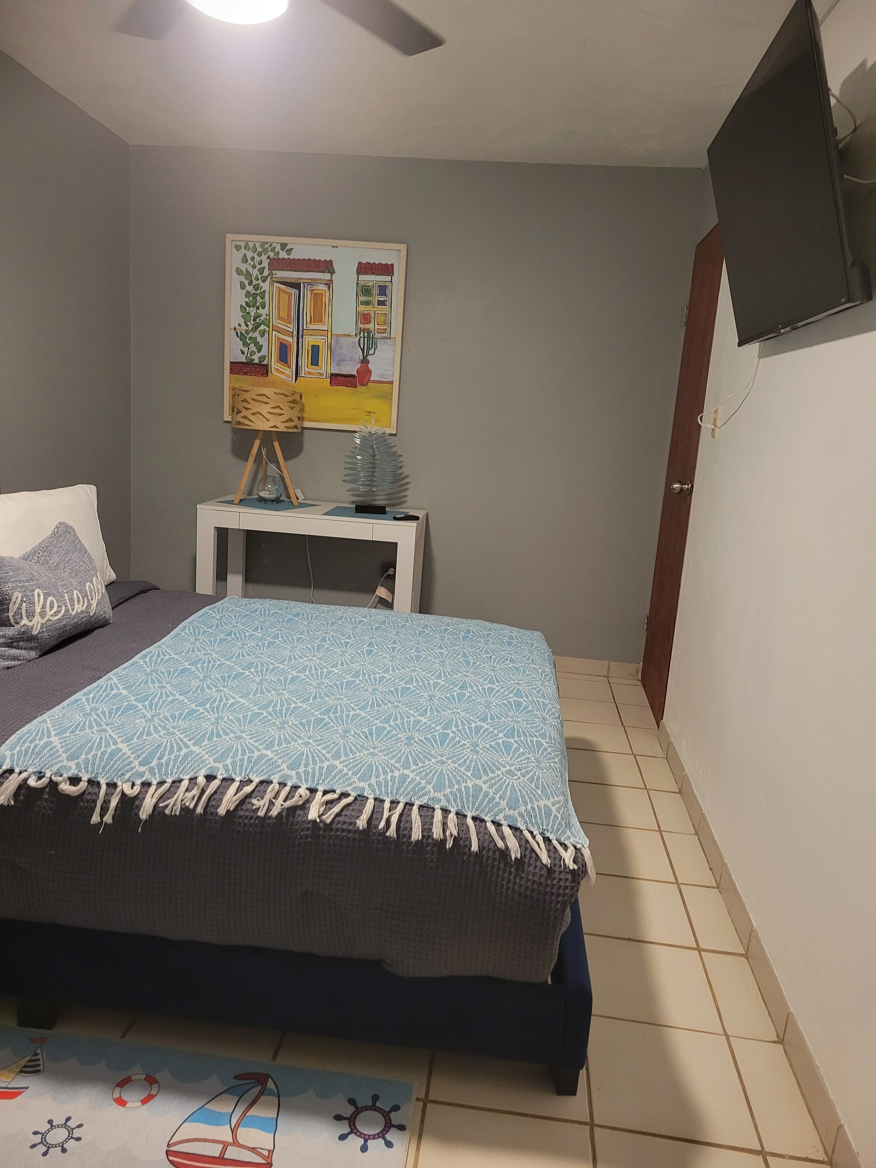 A bedroom with a bed, a small table with a plant, a painting on the wall, and a TV mounted on the wall.