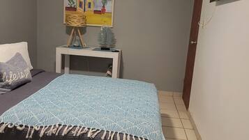 A bedroom with a bed, a small table with a plant, a painting on the wall, and a TV mounted on the wall.