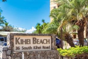 A sign for 'Kihei Beach' at 36 South Kihei Road, with a stone wall and palm trees in the background. - Kihei Beach 606 - Direct Ocean Front, Breathtaking Top Floor Views, Central AC! (Kihei)