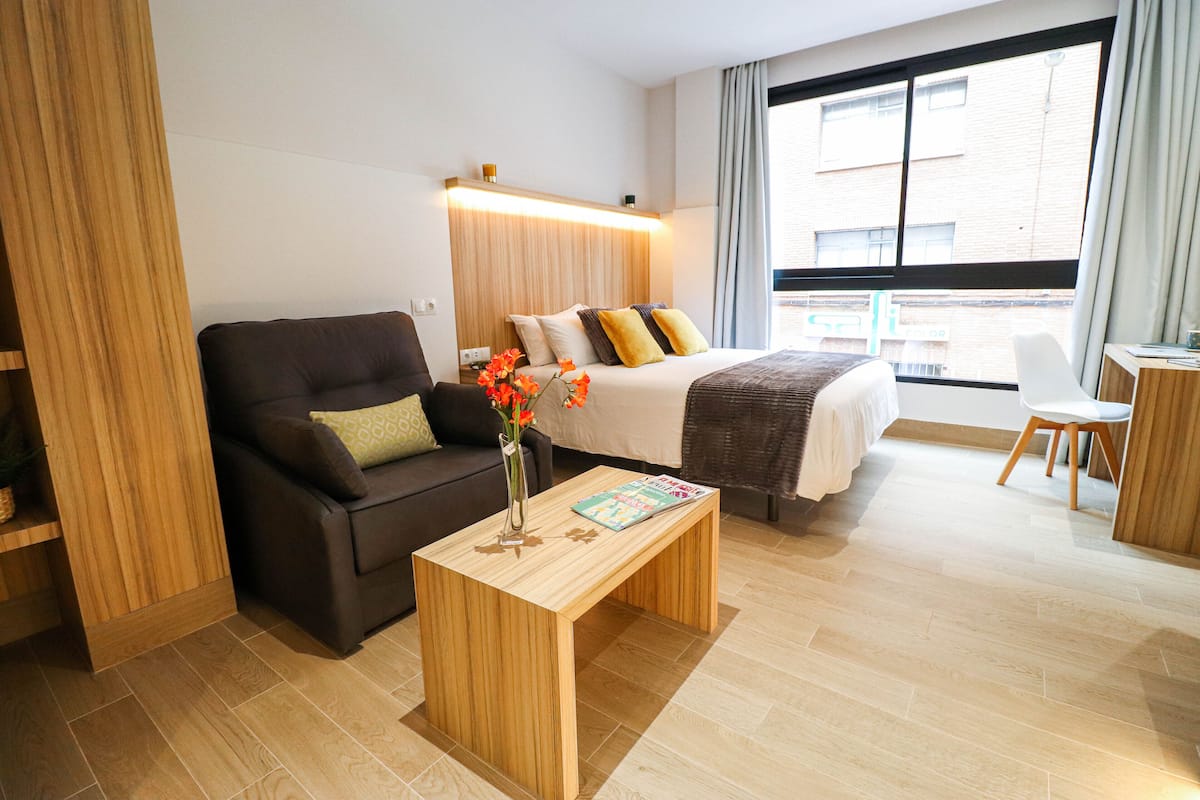 Una habitación de hotel moderna con un sofá, una mesa de centro de madera con una revista, una cama con almohadas y un ventanal con cortinas.