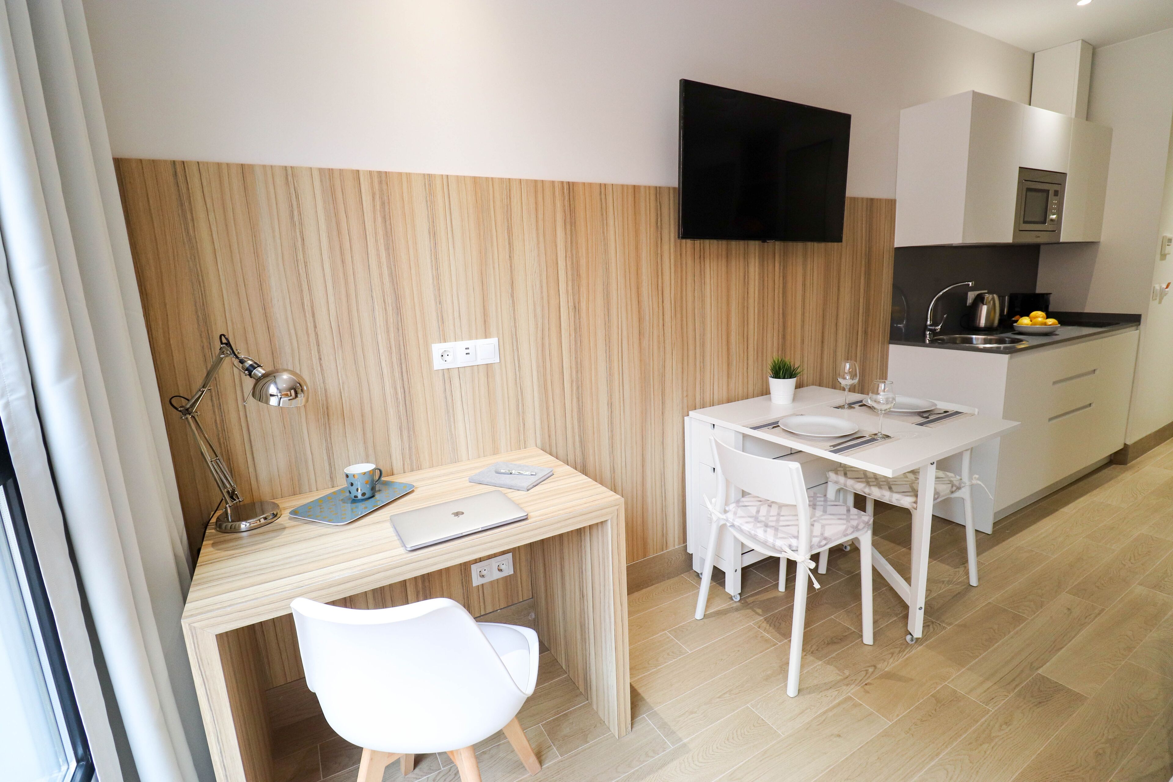 A compact kitchen with a dining area, a flat-screen TV mounted above the counter, and a small potted plant on the table.