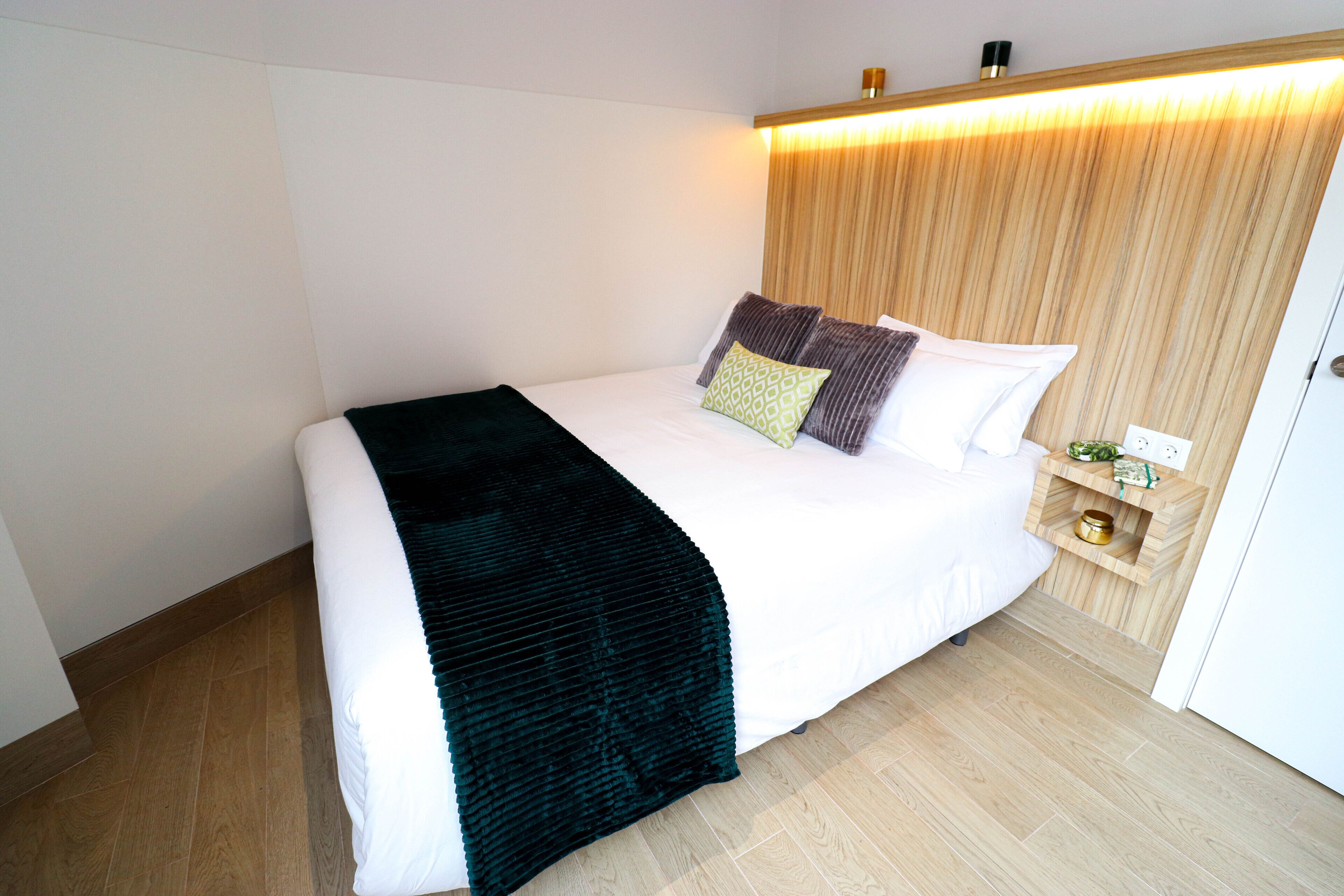A neatly made bed with a dark green throw blanket, a wooden headboard, and a bedside table with a small plant and a decorative item.