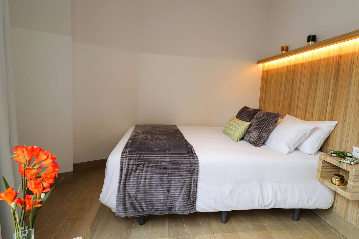 Una cama bien hecha con edredón blanco y manta gris oscuro, acompañada de varios almohadones. A la derecha de la cama, una mesita de noche de madera con una lámpara y algunos objetos.