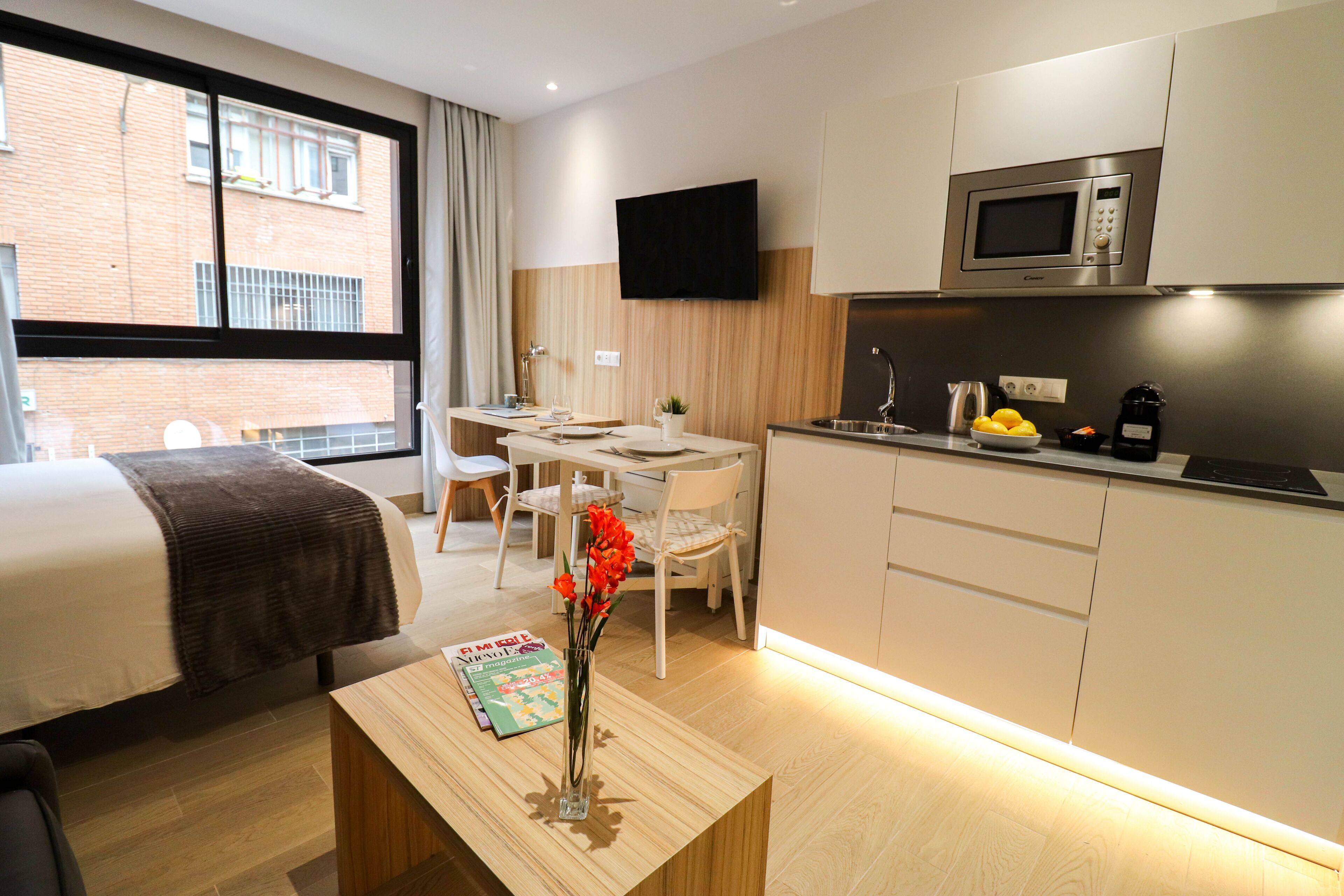 A modern hotel room with a bed, a desk, a chair, a small table with a magazine and a vase of flowers, a kitchenette with a microwave, and a TV on the wall.