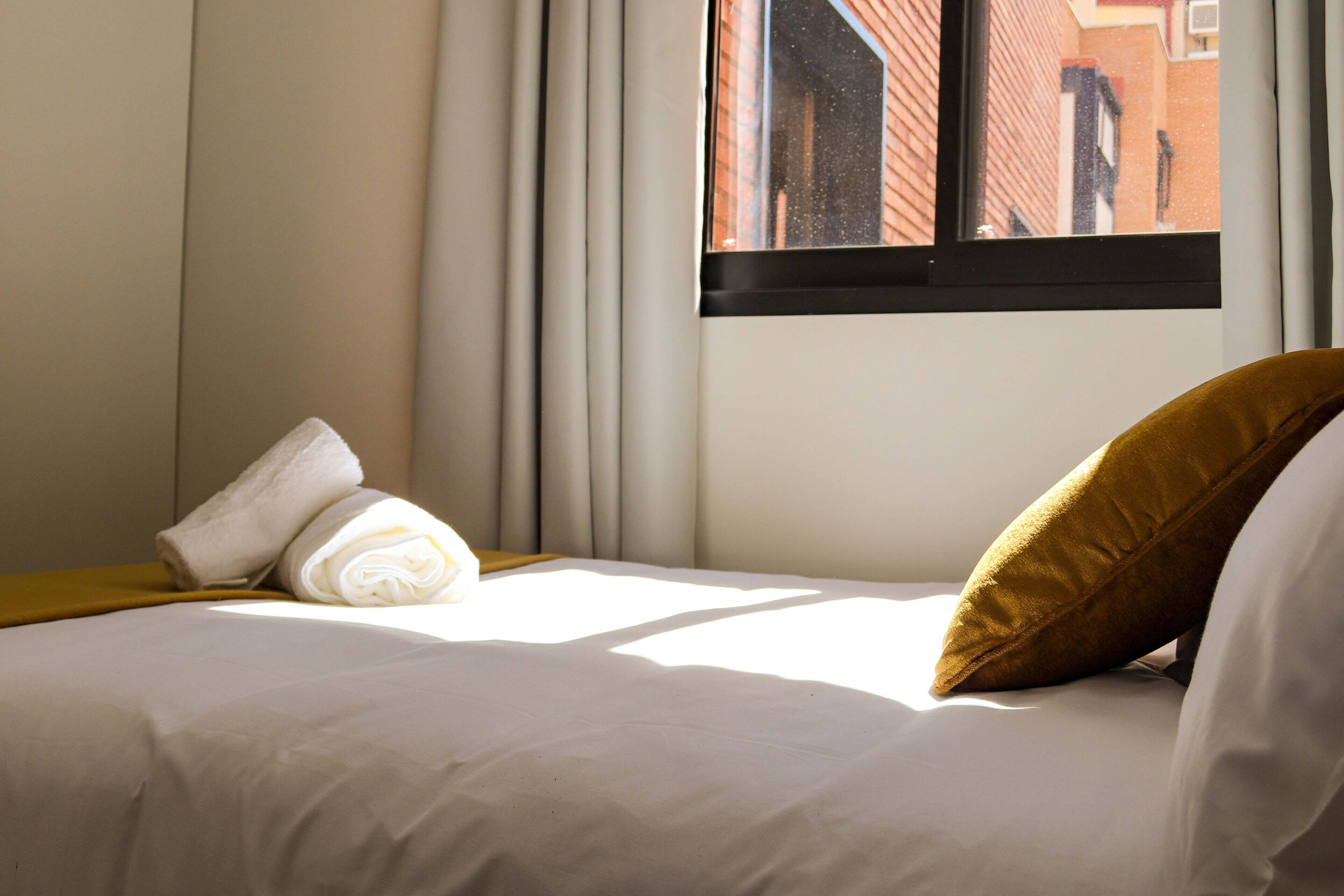 A neatly made bed with a rolled white towel and a brown pillow, a window with a view of a brick building, and a curtain partially drawn to the side.