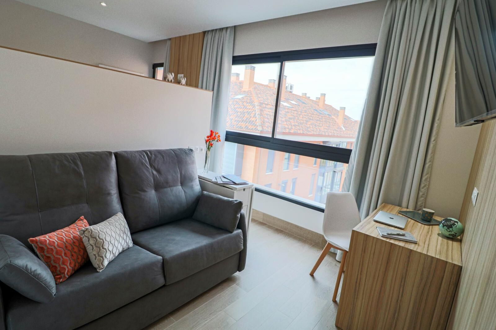 En moderne stue med en grå sofa, et lille skrivebord og et vindue med gardiner.