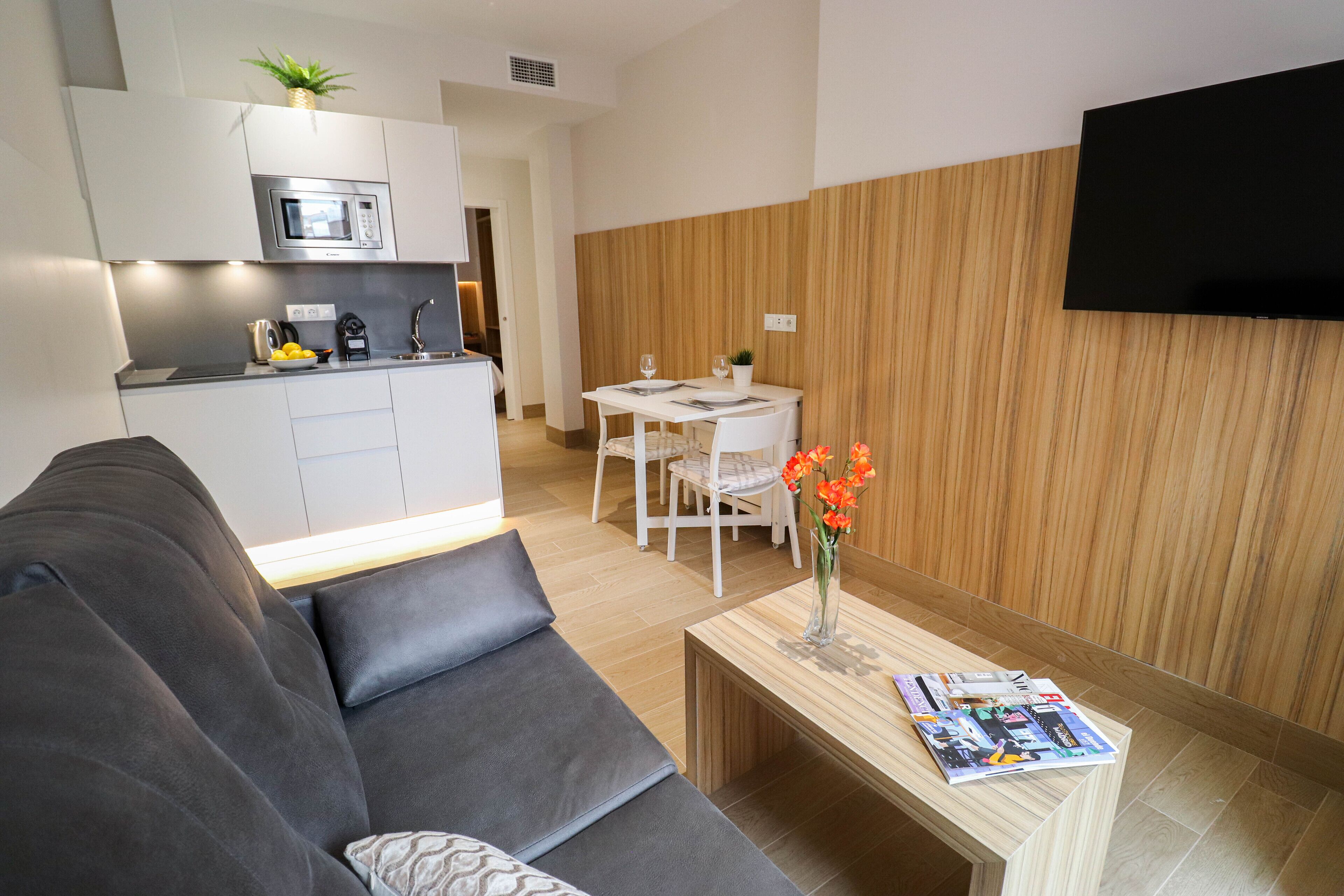 Una sala de estar moderna con un sofá gris, una mesa de centro de madera con revistas, y un comedor con una mesa y sillas blancas.