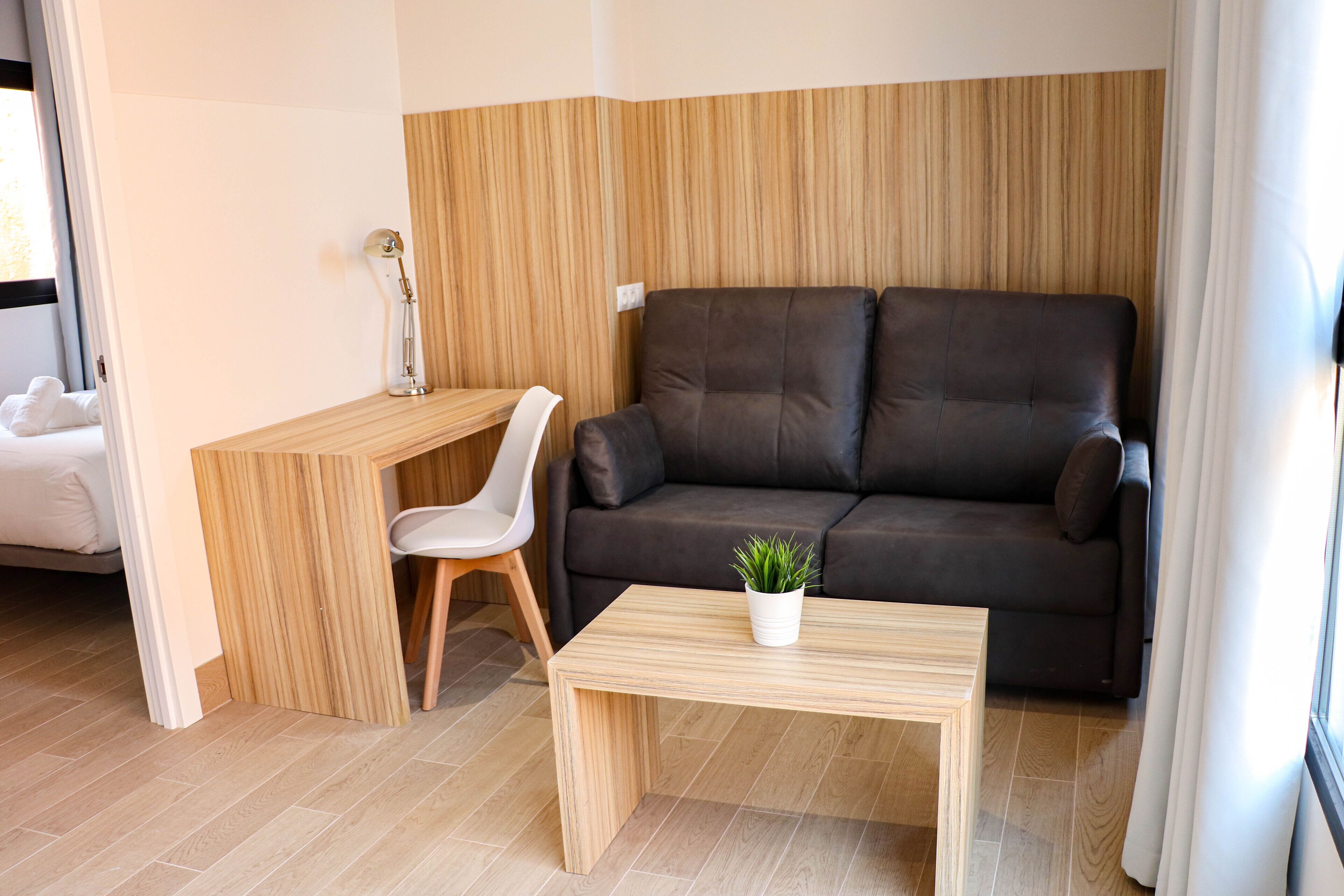 A modern living room with a brown leather sofa, a wooden coffee table with a plant, a white chair, and a desk with a lamp.