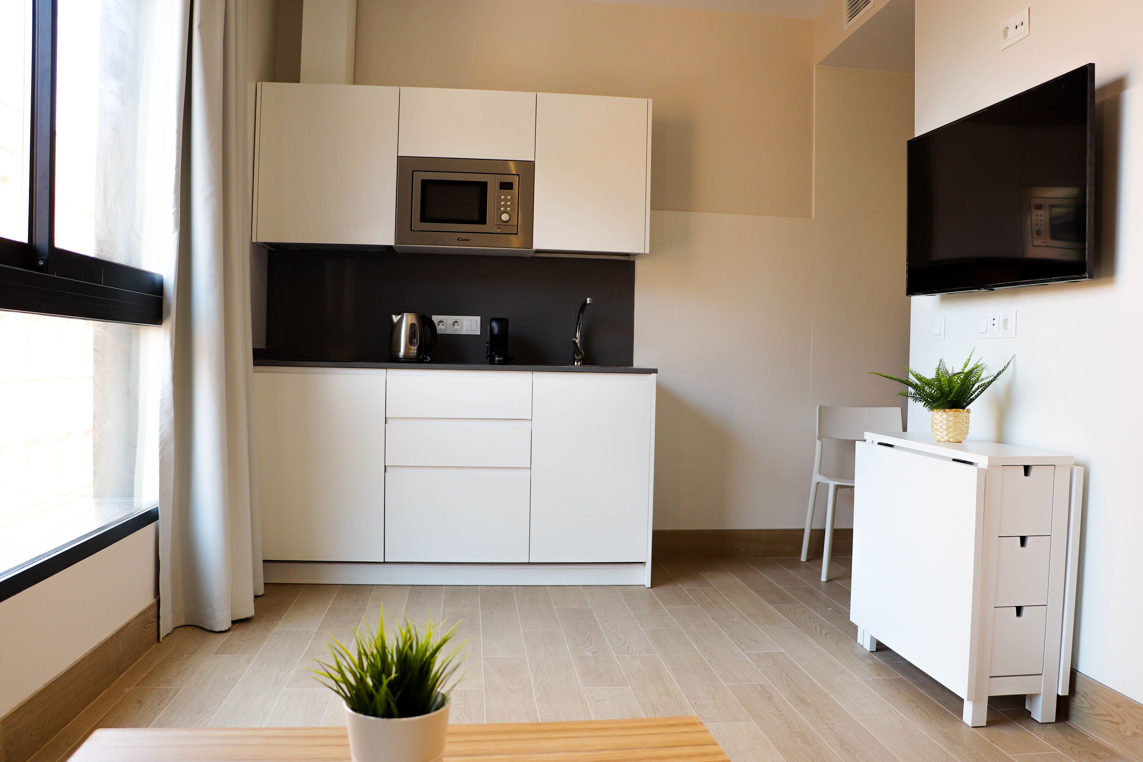 A modern kitchen with white cabinetry, a microwave, a kettle, and a small table with a plant.