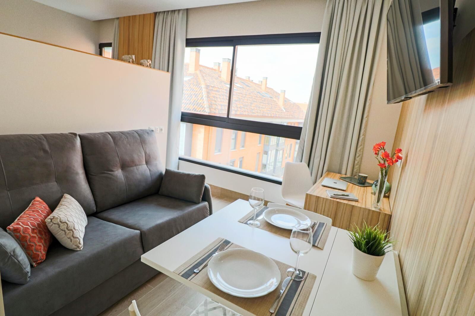 Un salon moderne avec un canapé gris, une table à manger dressée pour deux personnes et une grande fenêtre avec des rideaux.