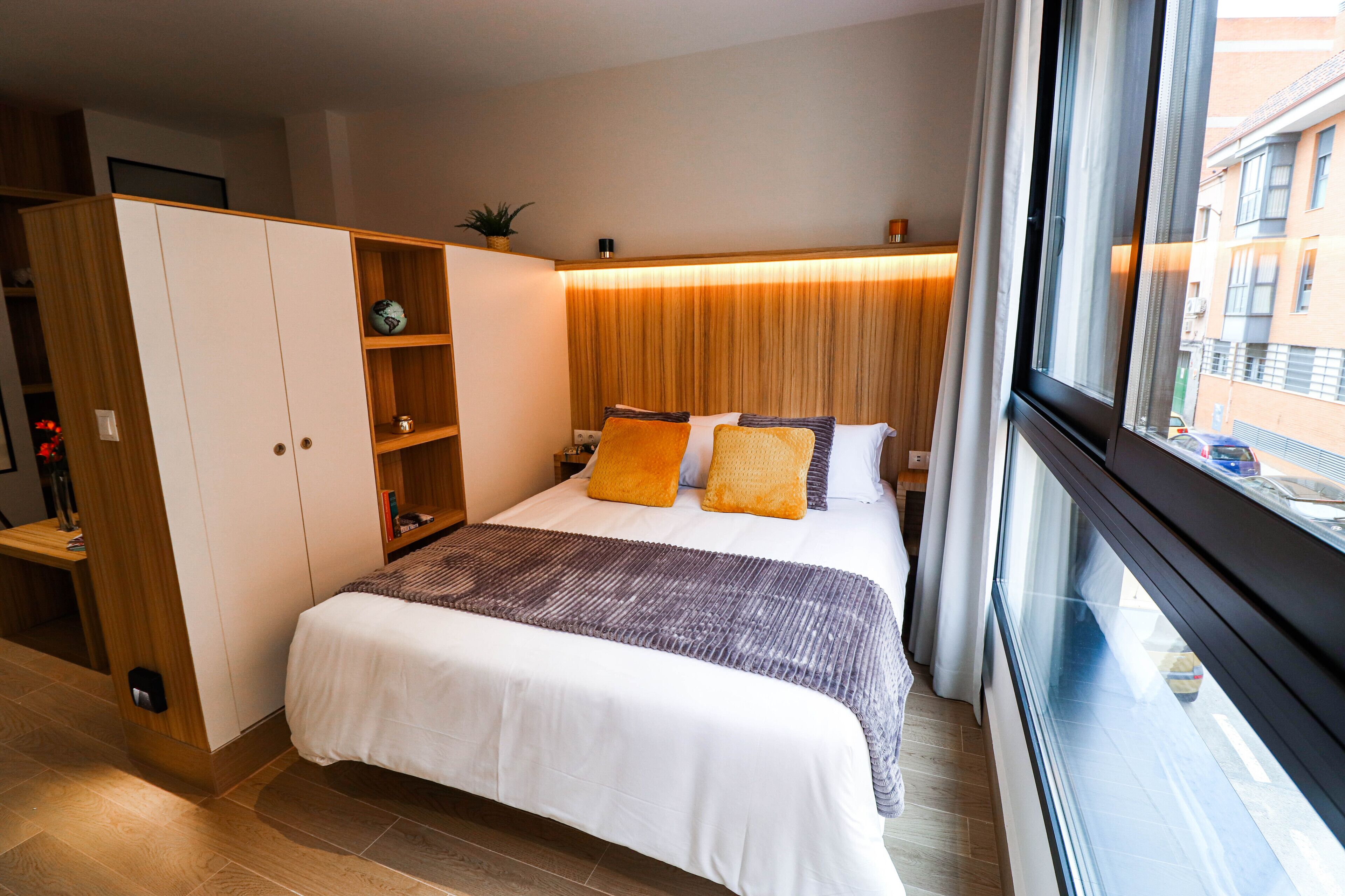 A modern hotel room with a large bed, wooden wardrobe, and a view of the city through a large window.