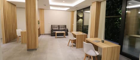 A modern hotel room with a dining area, a sofa, and a large window with a view of greenery.