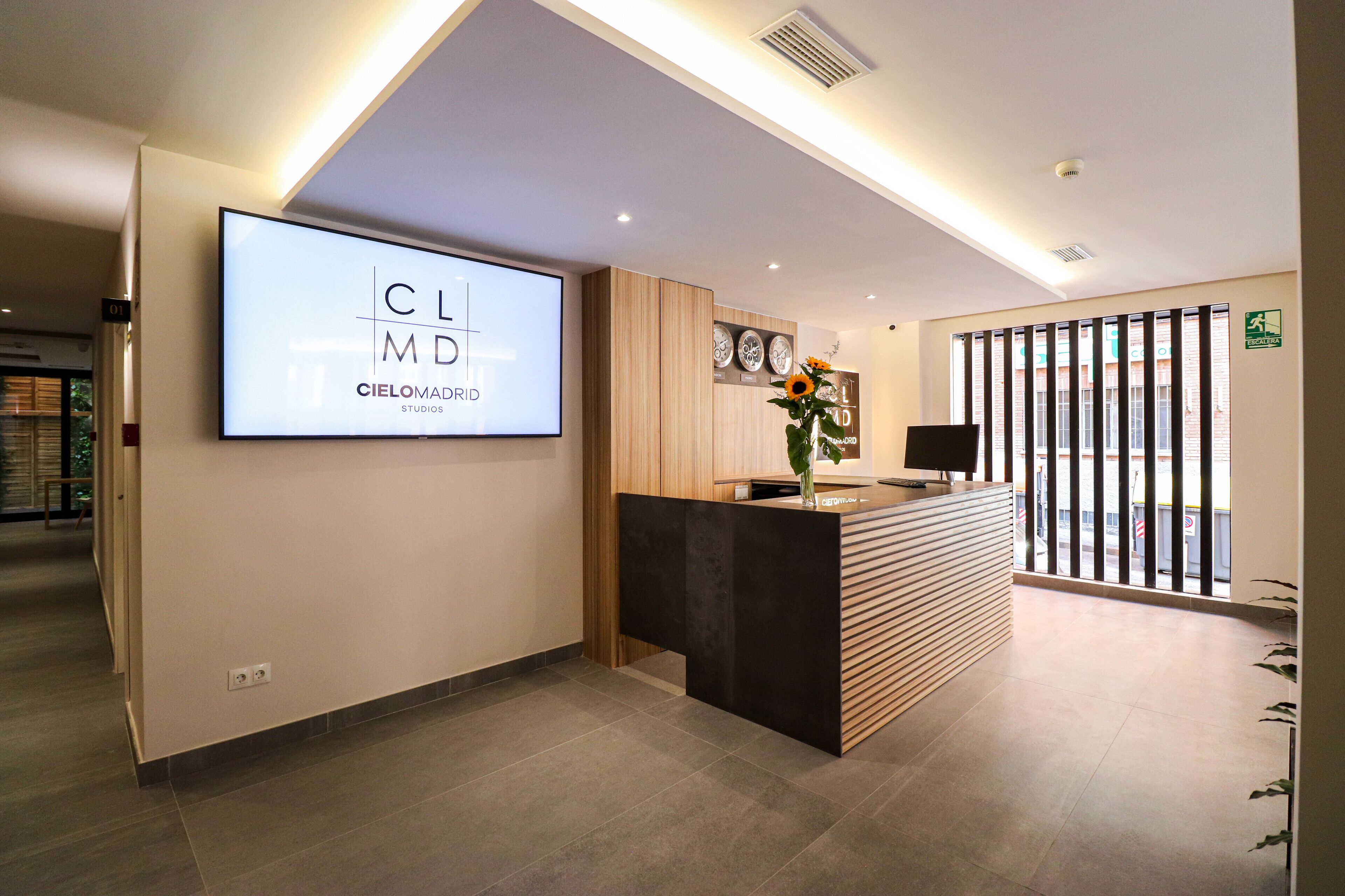 A modern reception area with a large digital display, a reception desk, and a view through a window with blinds.