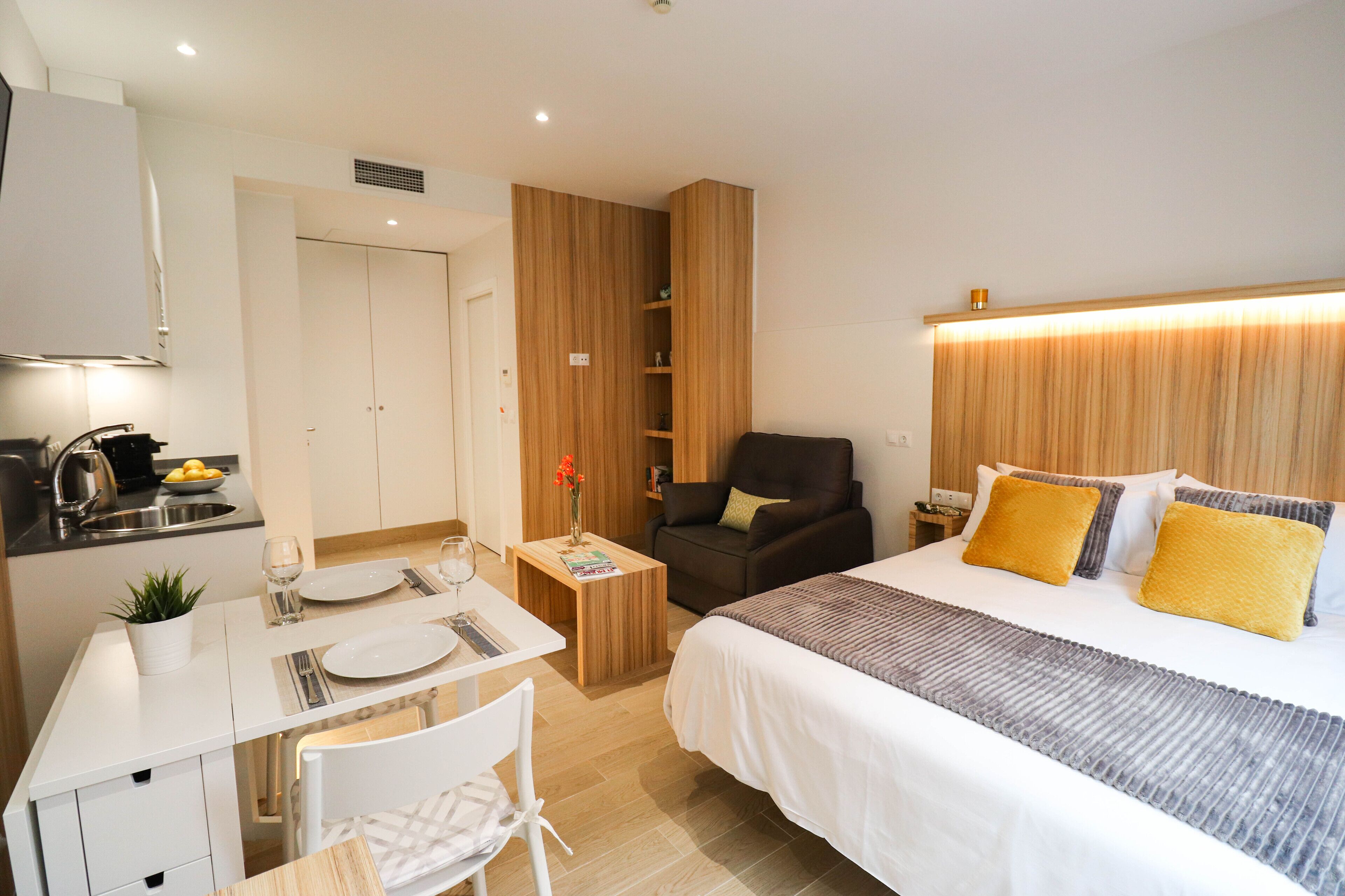 Una habitación de hotel compacta con cama, un escritorio pequeño con silla, kitchenette y armario.