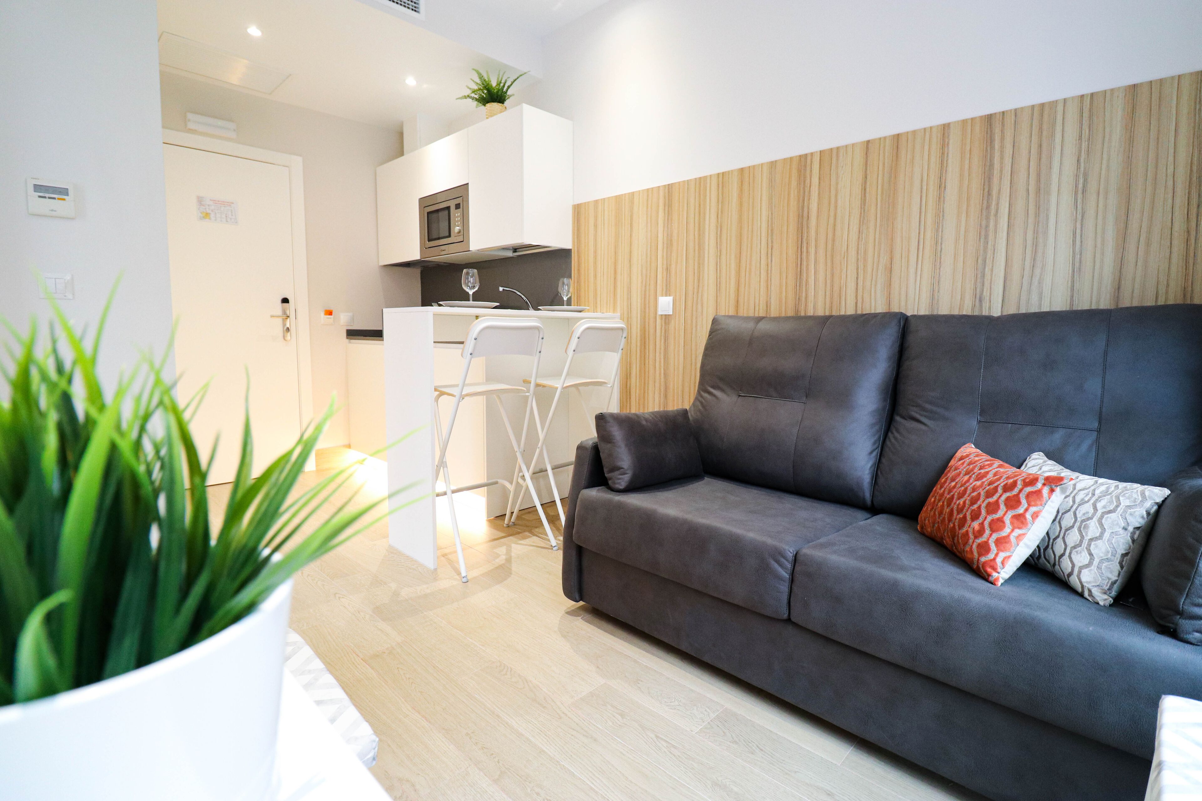 A modern living room with a grey sofa, two white chairs, a wooden accent wall, and a kitchen in the background.