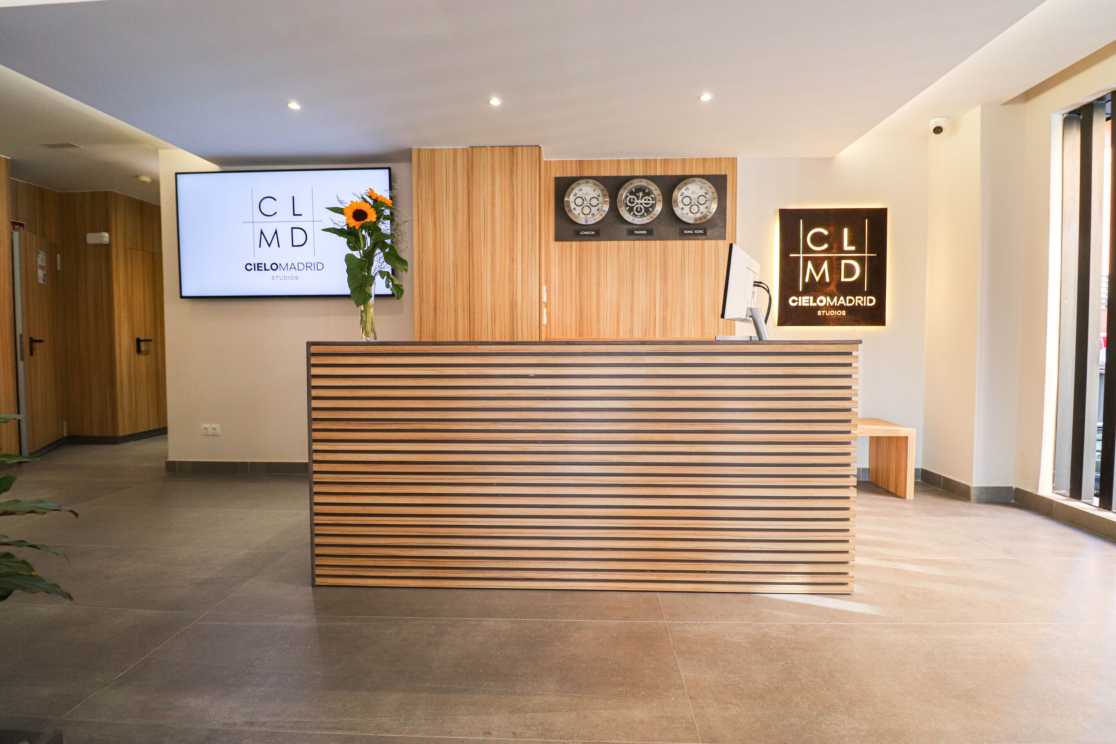 Taulell de recepció amb disseny de lamel·les de fusta, un vas amb flors i dos grans panells digitals amb el logotip 'CLMD CIELOMADRID STUDIO'.