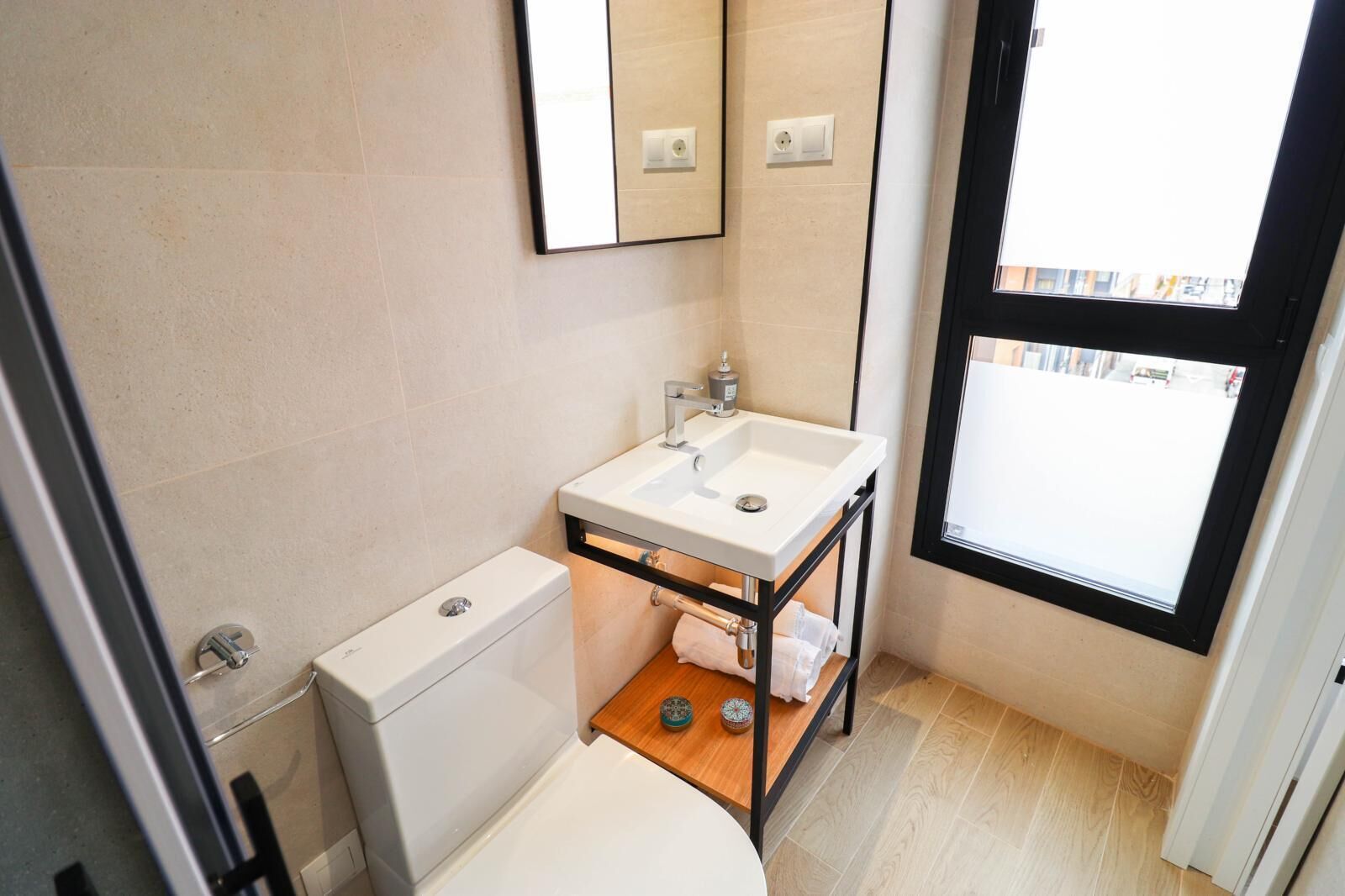 A compact bathroom with a white toilet, a sink on a black metal stand, and a window providing natural light.