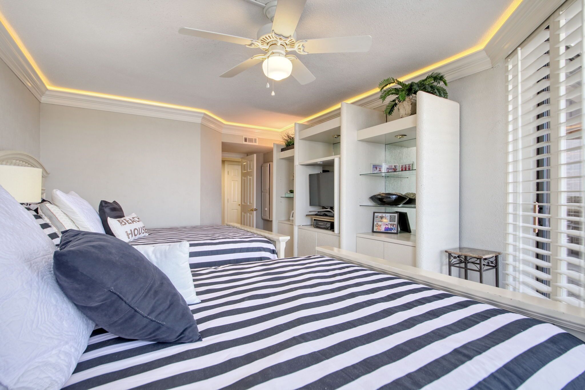 A hotel room with two beds, a ceiling fan, a built-in TV shelf, and a window with blinds.